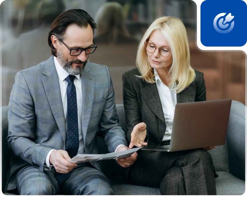 A business meeting between two professionals seated on a gray couch, discussing documents and using a laptop.