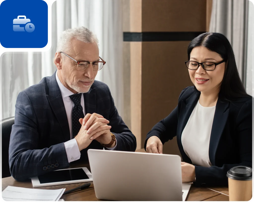 Two professionals engaged in a business discussion at a modern office table, with a laptop, tablet, documents, and coffee cup.