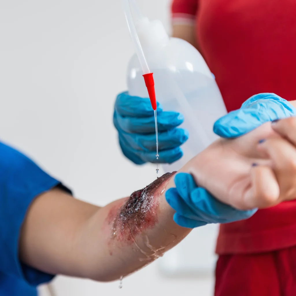 A person sitting indoors is receiving an injection from a healthcare professional wearing a medical glove. The focus is on the wrist where the needle is being administered. Medical equipment is visible nearby.