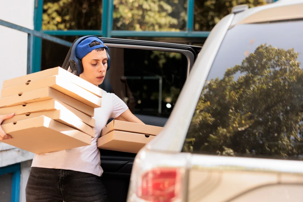 A person carrying multiple pizza boxes while leaning against a car, with trees visible in the background through a window.