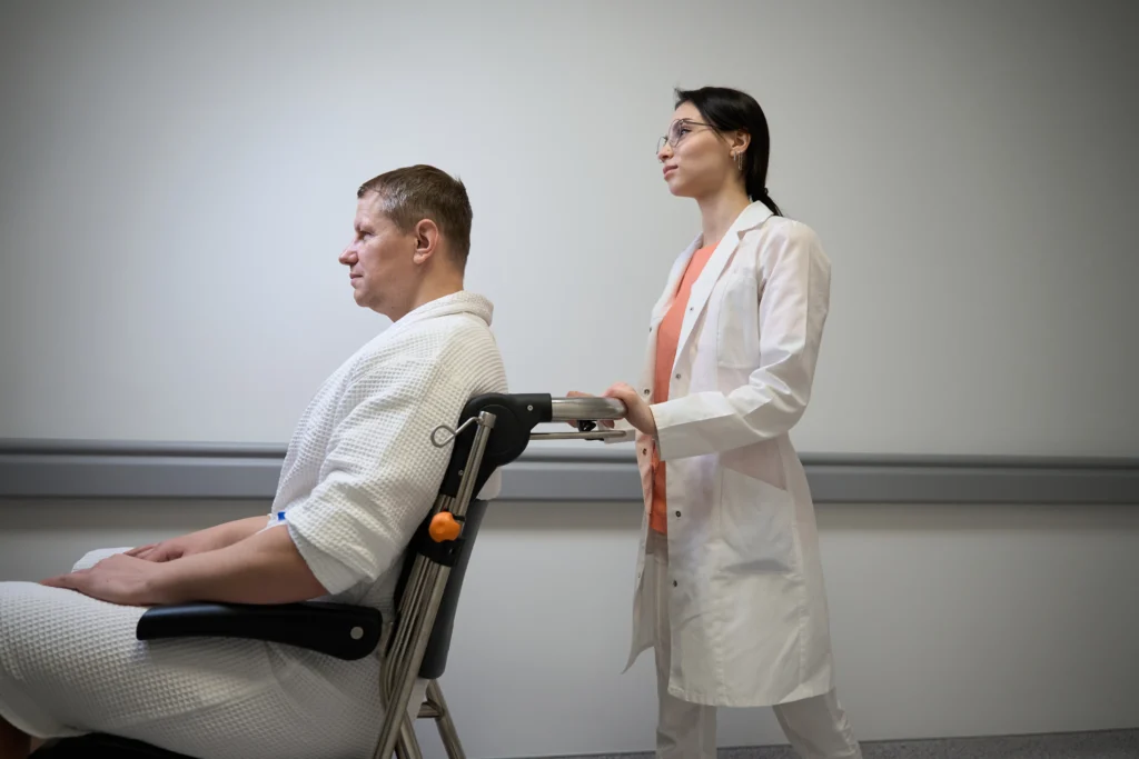 A healthcare professional in a white lab coat pushes a patient in a hospital gown on a wheelchair through a clinical hallway.