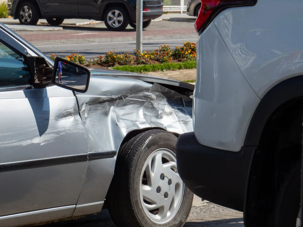A silver car with significant damage to its driver's side collides with a white SUV, parked on a sunny street.
