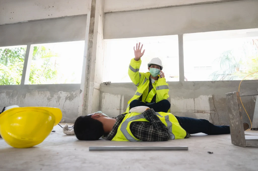 A person in a safety jacket is sitting on the floor, making a call while another individual lies unconscious nearby, with construction tools around.
