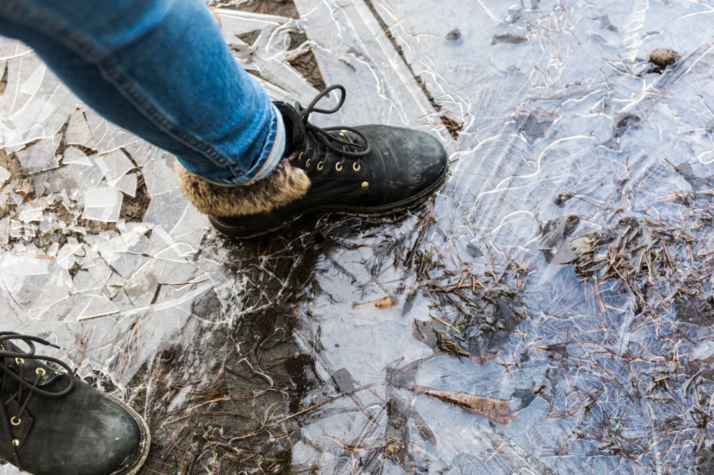 A close-up of black boots standing on thin ice, with cracks and pine needles visible in the frozen surface beneath.