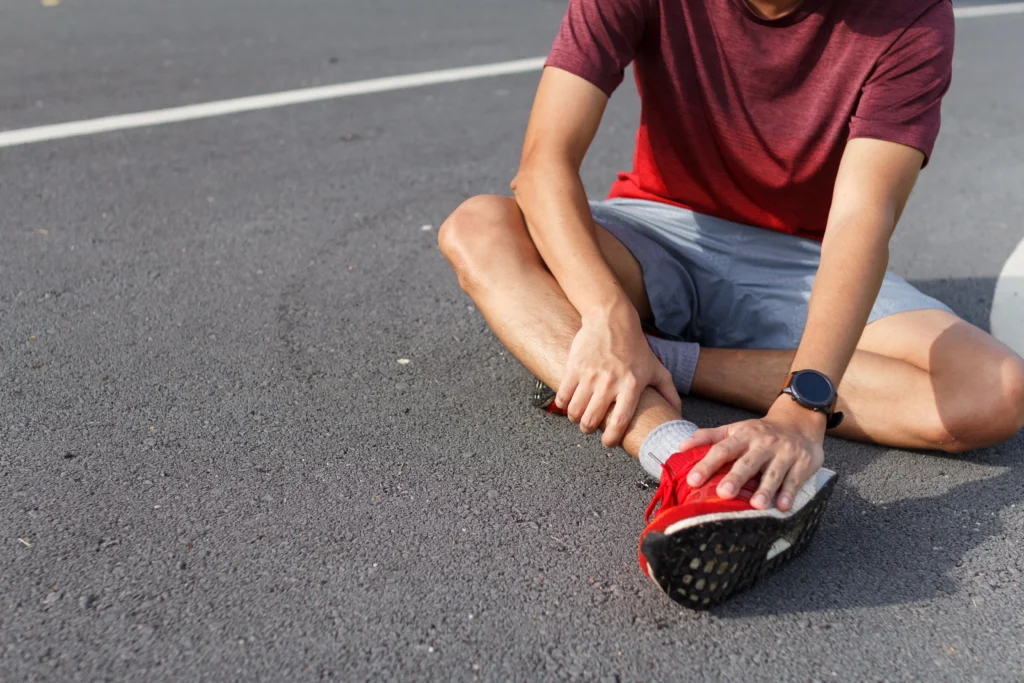 A young man in a red shirt is sitting on the pavement, holding his ankle in discomfort after an apparent injury.