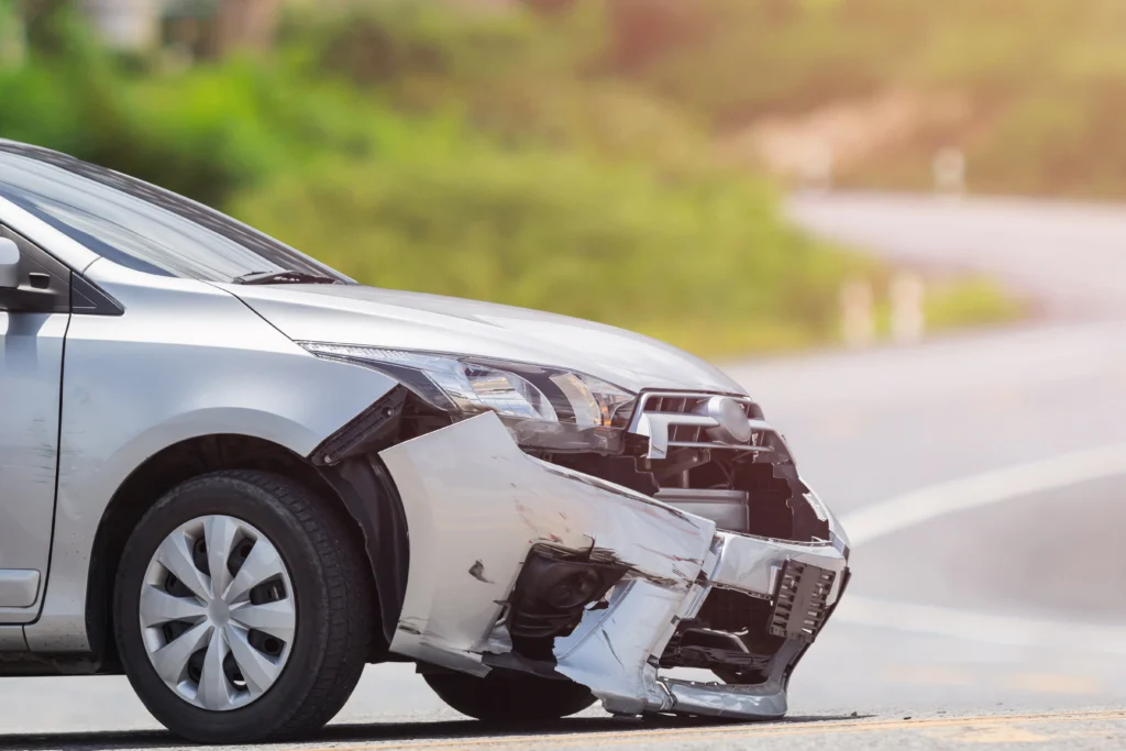 A silver car with a severely damaged front bumper and fender, parked on the roadside with blurred greenery in the background.