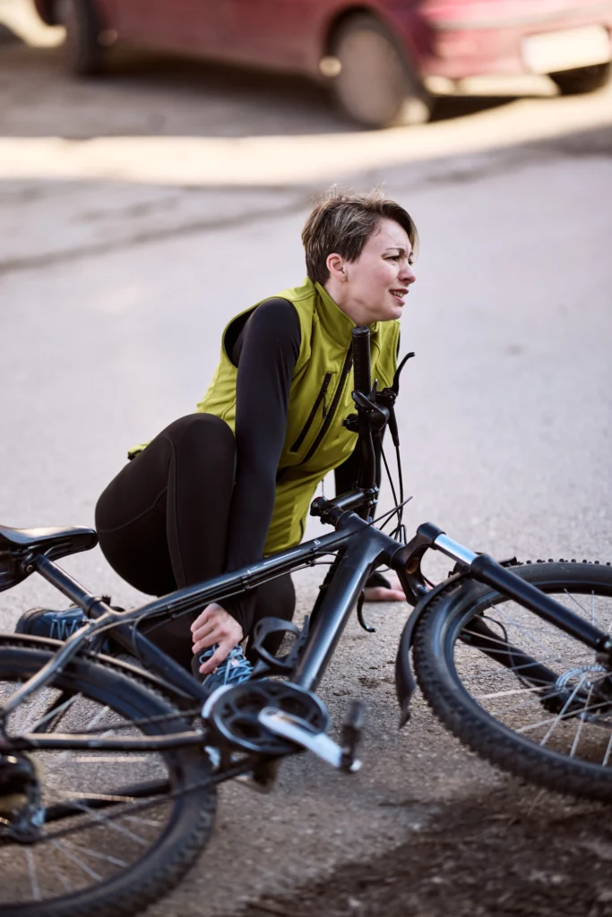 A person in a green vest and black leggings crouches beside a fallen mountain bike on a gravel surface.