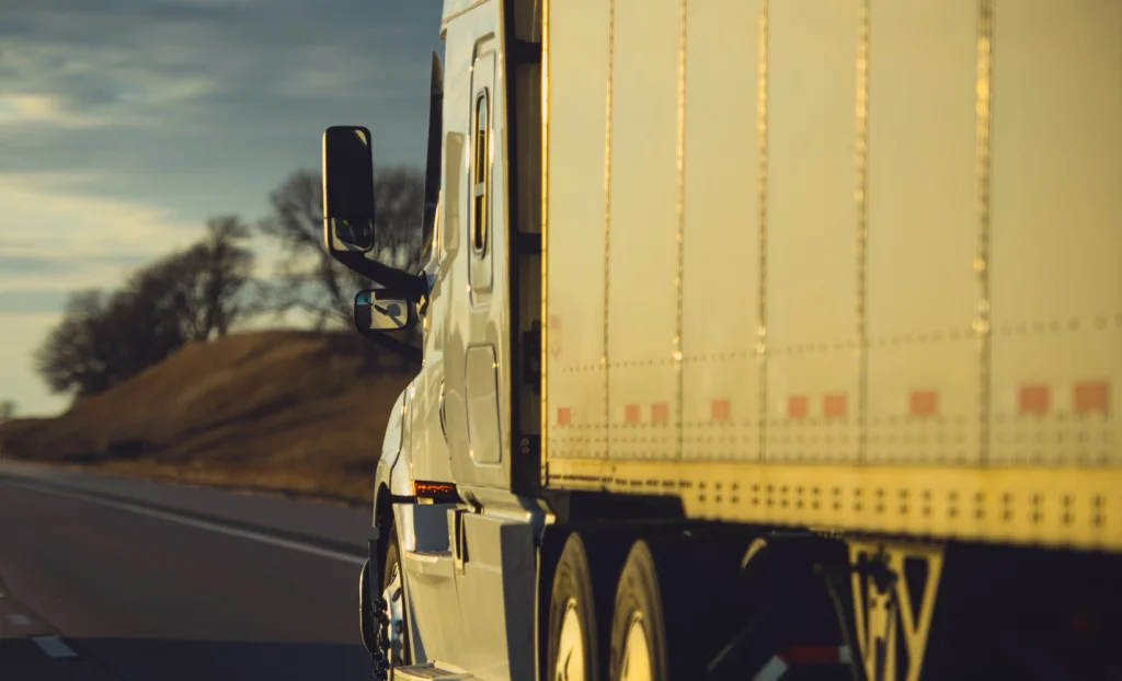 A close-up of a white semi-truck on the highway, showcasing its side mirror and trailer against a golden sunset backdrop.