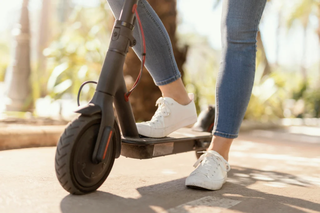 A close-up of a person's foot on a black electric scooter, wearing white sneakers, with a sunny path and greenery in the background.