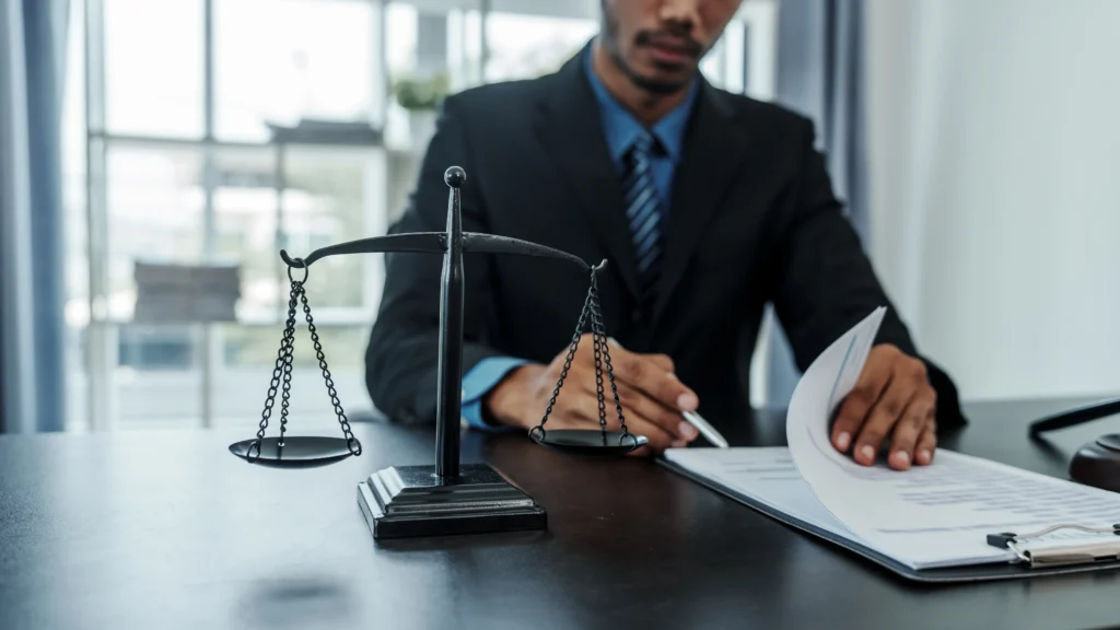 A lawyer in a suit is seated at a desk, with a balance scale and documents in front, emphasizing a legal setting.